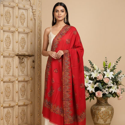 Woman wearing a red shawl with intricate patterns, standing next to a decorative vase with flowers and an ornate wooden door.