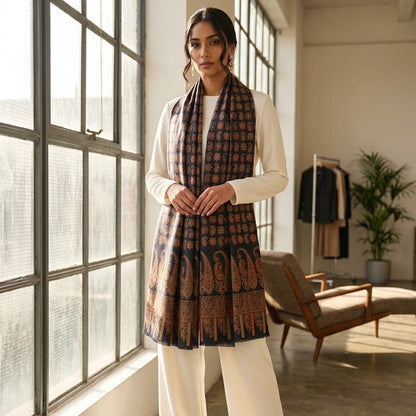 A woman models a traditional Indian outfit consisting of a white kurta, white wide-leg trousers, and a dark brown patterned dupatta with gold motifs, standing in a bright, modern loft with large windows.