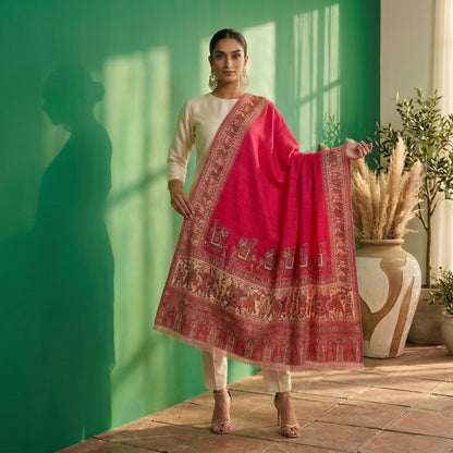 A woman models a traditional outfit consisting of a cream kurta and trousers paired with a vibrant pink dupatta featuring intricate gold and red border patterns, set against a green wall with potted plants.