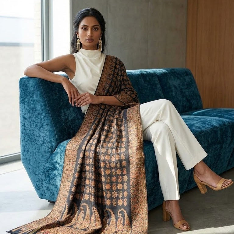 A woman sits on a teal velvet sofa wearing a white sleeveless top and white pants, with a large patterned brown and black shawl draped over her body in a modern indoor setting.