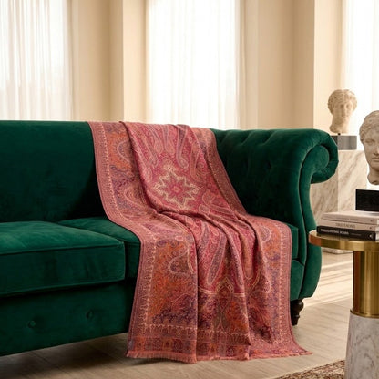 A pink and red paisley patterned throw blanket is draped over a deep green velvet sofa in a beautifully lit, elegant living room with cream walls, arched windows, and a marble side table.