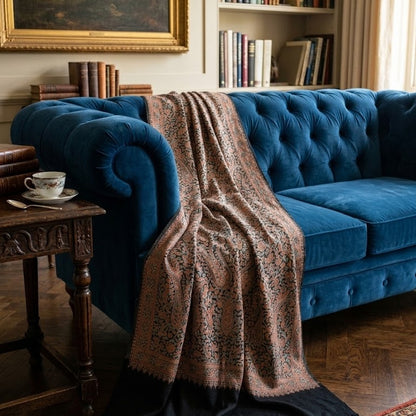 A blue velvet tufted sofa in a cozy library setting, draped with a beige and black patterned shawl. A wooden side table with a teacup sits to the left, and bookshelves are visible in the background.