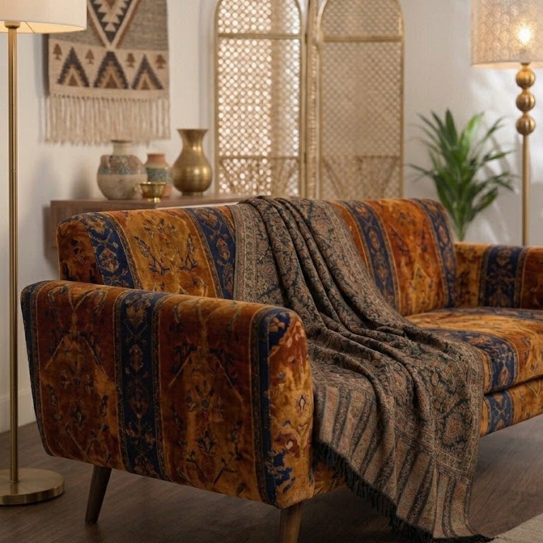 A mid-century modern loveseat sofa upholstered in a richly patterned rust-orange and navy blue velvet fabric, styled with a matching patterned throw blanket draped over the seat. The sofa is set in a warm, bohemian-style living room with wooden floors, a tall floor lamp, a patterned wall hanging, and a rattan screen.