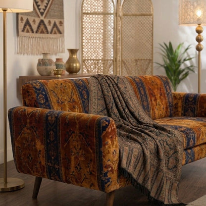 A mid-century modern loveseat sofa upholstered in a richly patterned rust-orange and navy blue velvet fabric, styled with a matching patterned throw blanket draped over the seat. The sofa is set in a warm, bohemian-style living room with wooden floors, a tall floor lamp, a patterned wall hanging, and a rattan screen.