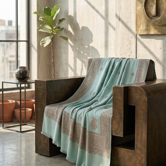 A draped mint green and beige patterned throw blanket is shown on a dark brown wooden armchair in a modern, sunlit concrete room with a large window and plant.