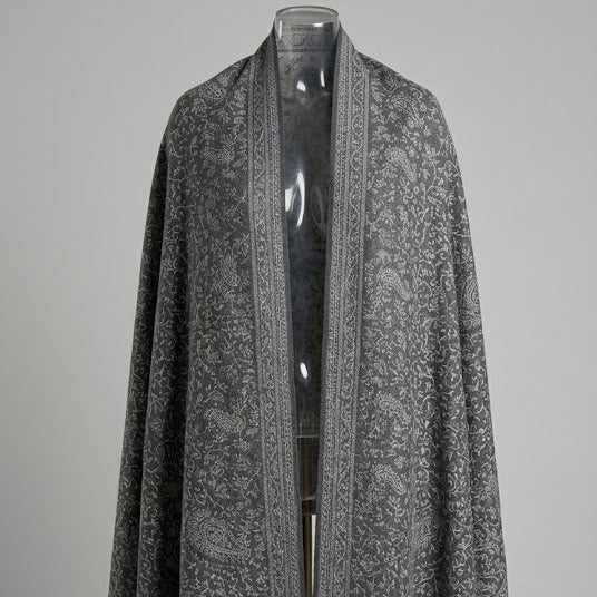A gray and black patterned shawl draped over a clear mannequin torso, featuring intricate paisley and floral motifs with a fringed hem, set against a neutral gray background.