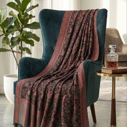 A dark teal velvet armchair in a bright living room, draped with a patterned throw blanket featuring rust red borders and a dark floral motif.