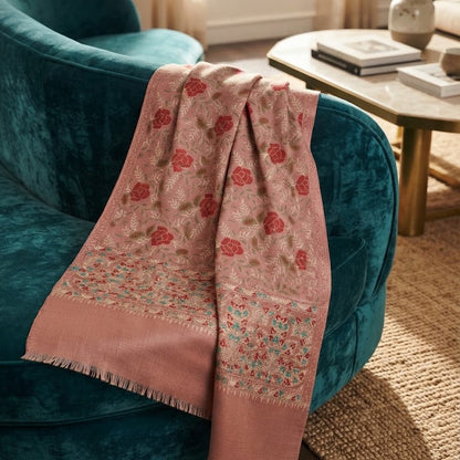 A dusty pink woven throw blanket with red and teal floral motifs draped over a teal velvet armchair in a warm, sunlit living room.
