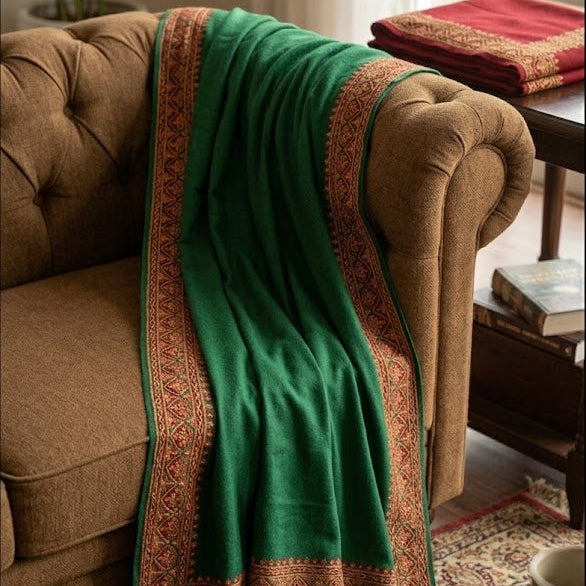 A green shawl with ornate gold and red woven borders draped over a brown tufted armchair in a cozy living room setting.
