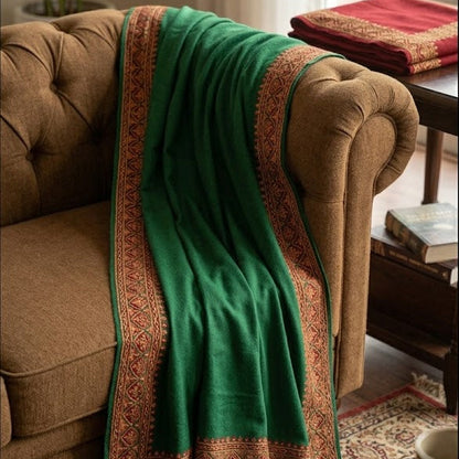 A green shawl with ornate gold and red woven borders draped over a brown tufted armchair in a cozy living room setting.