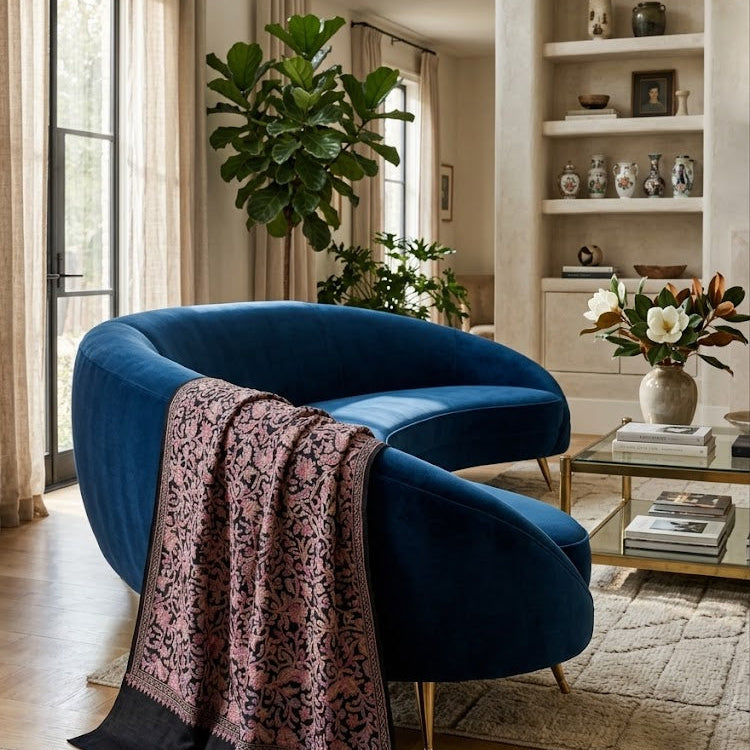 A curved blue velvet armchair in a bright, modern living room, styled with a pink and black patterned throw blanket draped over the seat.
