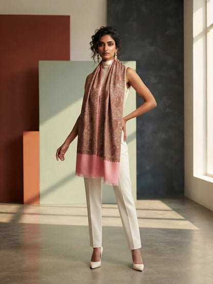 A woman models a draped scarf with a brown and pink paisley pattern and a solid pink border, styled with white trousers and white heels in a modern studio setting with geometric panels.
