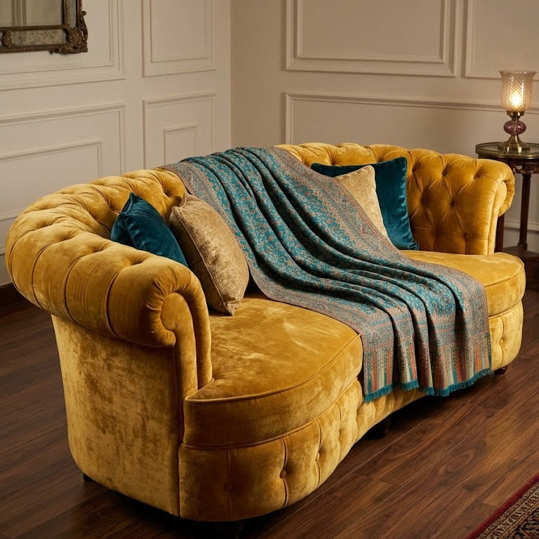 A curved mustard yellow velvet chaise lounge sofa styled with teal and beige throw pillows and a blue patterned throw blanket, set in a classic room with white paneled walls and dark wood floors.