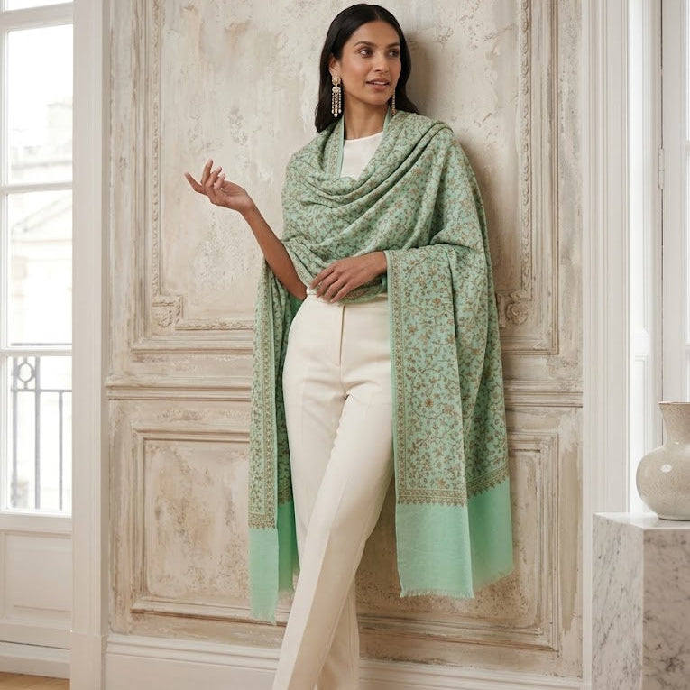 A woman models a sage green patterned shawl with mint green borders, draped over her shoulders, paired with white pants in an elegant room with ornate wall paneling.