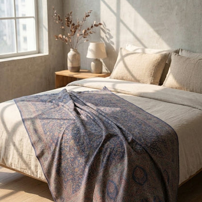 A patterned kani jamawar shawl draped over a bed in a sunlit bedroom. The blanket has a muted blue and dusty rose traditional pattern on a taupe base, while the bed is dressed in neutral beige linens with matching pillows.