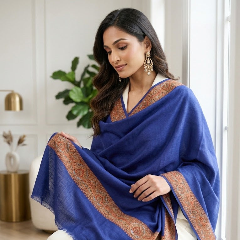 A woman seated indoors wearing a royal blue shawl with ornate gold and beige border detailing, paired with cream trousers. The setting is a bright, modern room with neutral walls, a plant, and gold-toned decor.