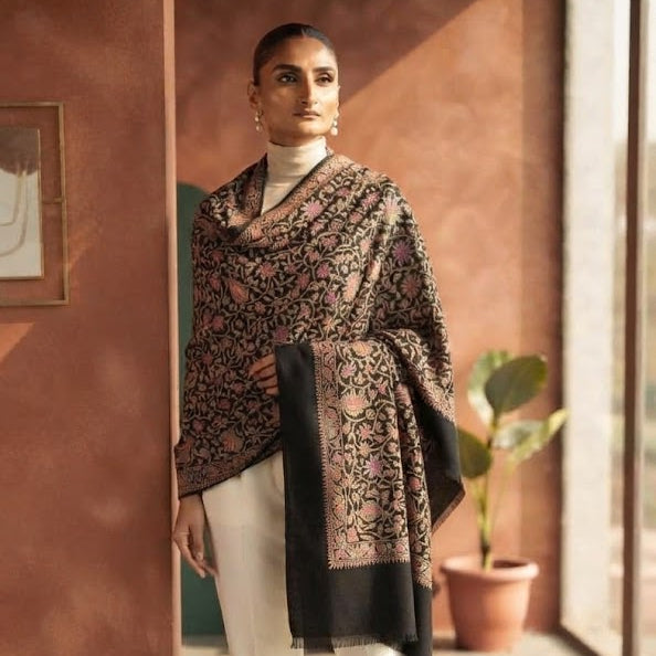 A woman models a black and brown patterned shawl draped over her shoulders, paired with white trousers and beige heels, standing in a warm-toned indoor corridor.