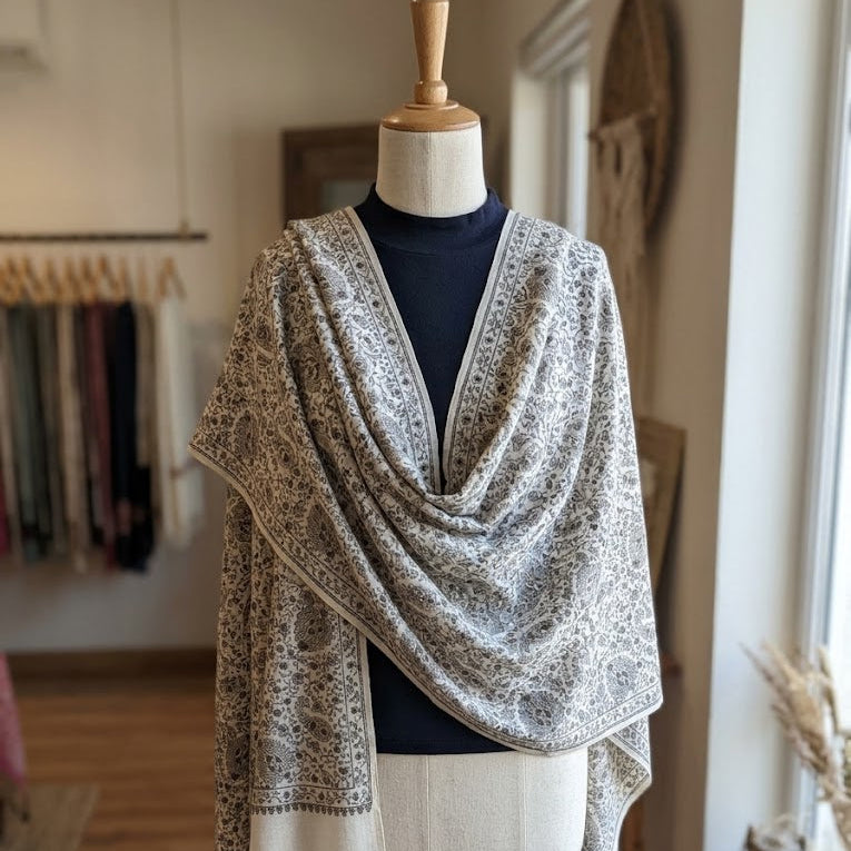 A patterned shawl with fringed edges draped over a dress form mannequin in a boutique setting.