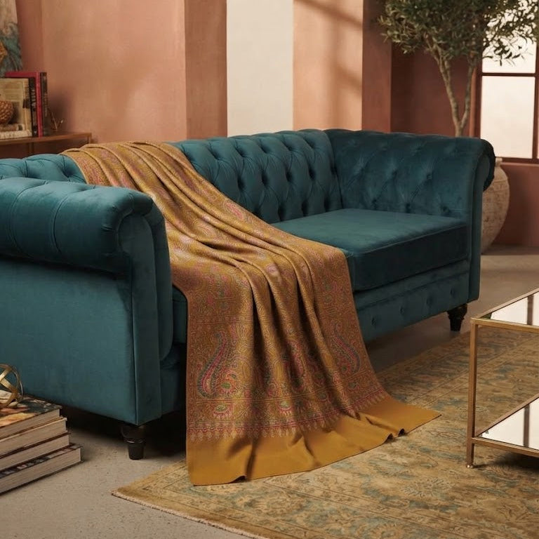 A teal velvet tufted loveseat sofa in a warm, rustic room setting, draped with a golden-brown patterned throw blanket.