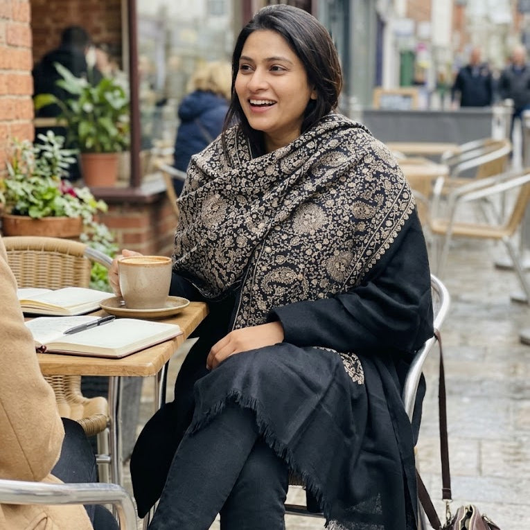 A woman sitting at an outdoor cafe table wearing a black and beige patterned shawl over a black outfit, with a coffee cup on the table in front of her.