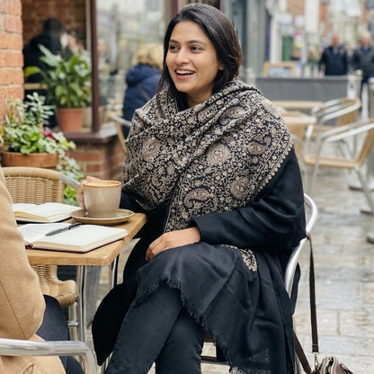 A woman sitting at an outdoor cafe table wearing a black and beige patterned shawl over a black outfit, with a coffee cup on the table in front of her.