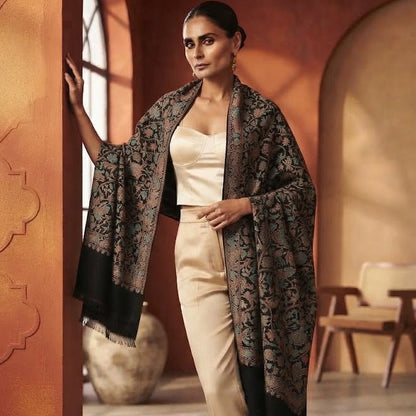 A woman models a black and beige patterned shawl draped over her shoulders, paired with a cream top and beige trousers, in a warm-toned indoor setting with arched walls.
