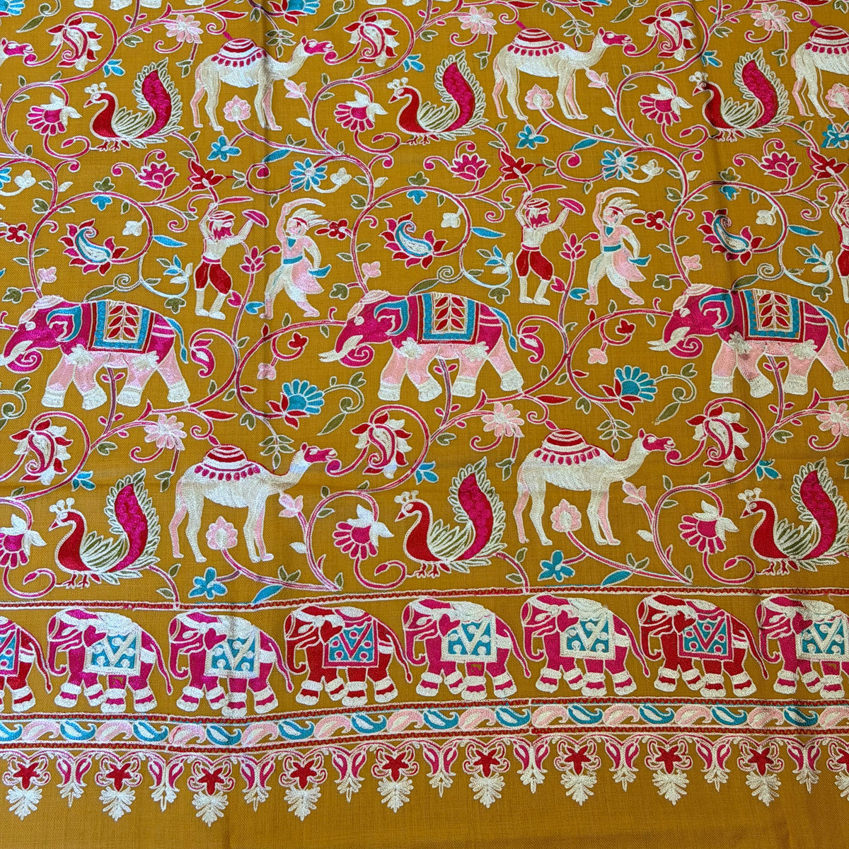 A close-up of a mustard yellow fabric with an intricate hand-block printed pattern featuring white elephants, pink and red birds, and a decorative border on the right side.
