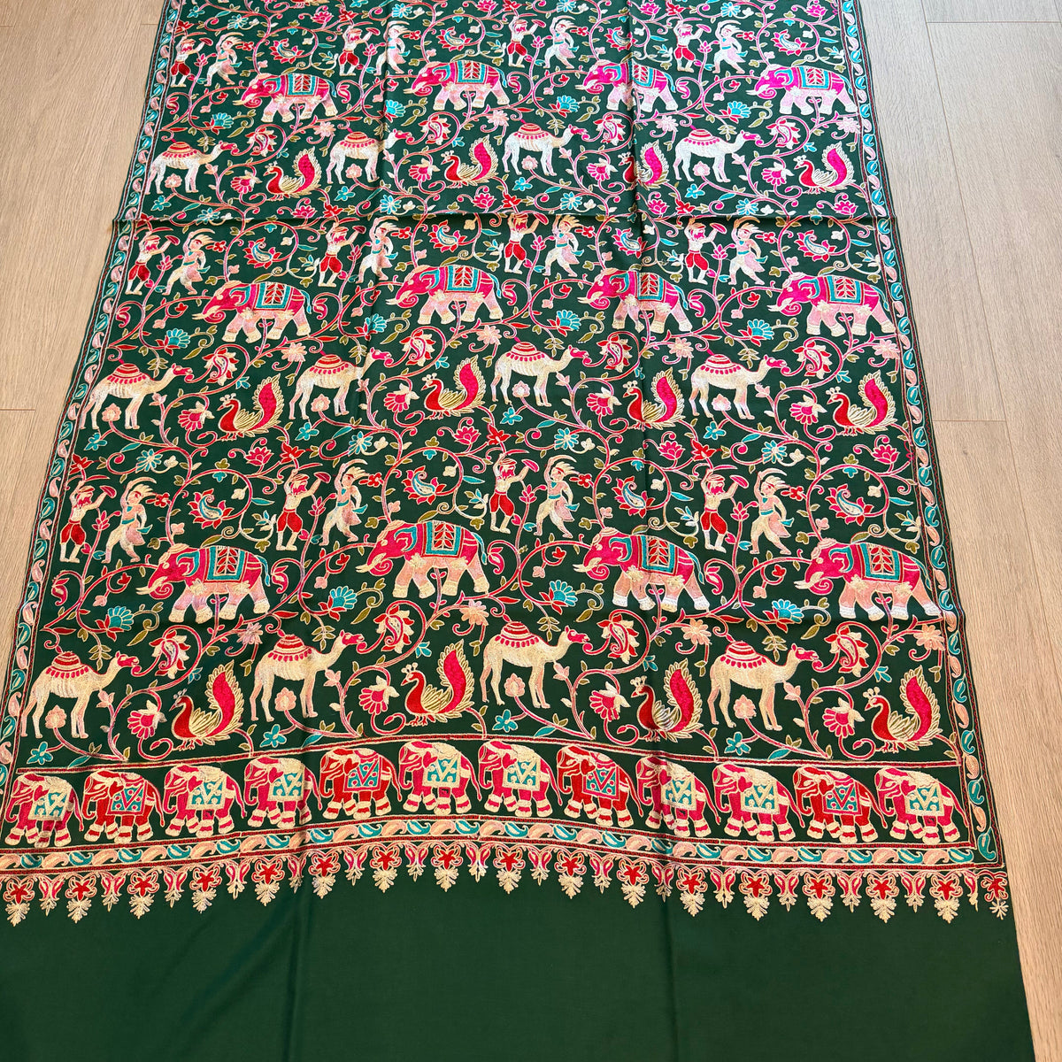 A dark green silk saree or fabric panel featuring intricate hand-embroidered motifs of elephants, deer, and birds in pink, red, white, and gold, laid flat on a light wood floor.