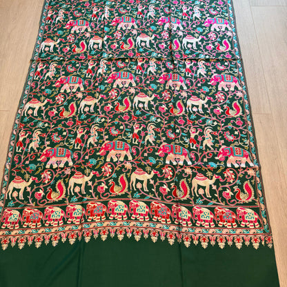 A dark green silk saree or fabric panel featuring intricate hand-embroidered motifs of elephants, deer, and birds in pink, red, white, and gold, laid flat on a light wood floor.