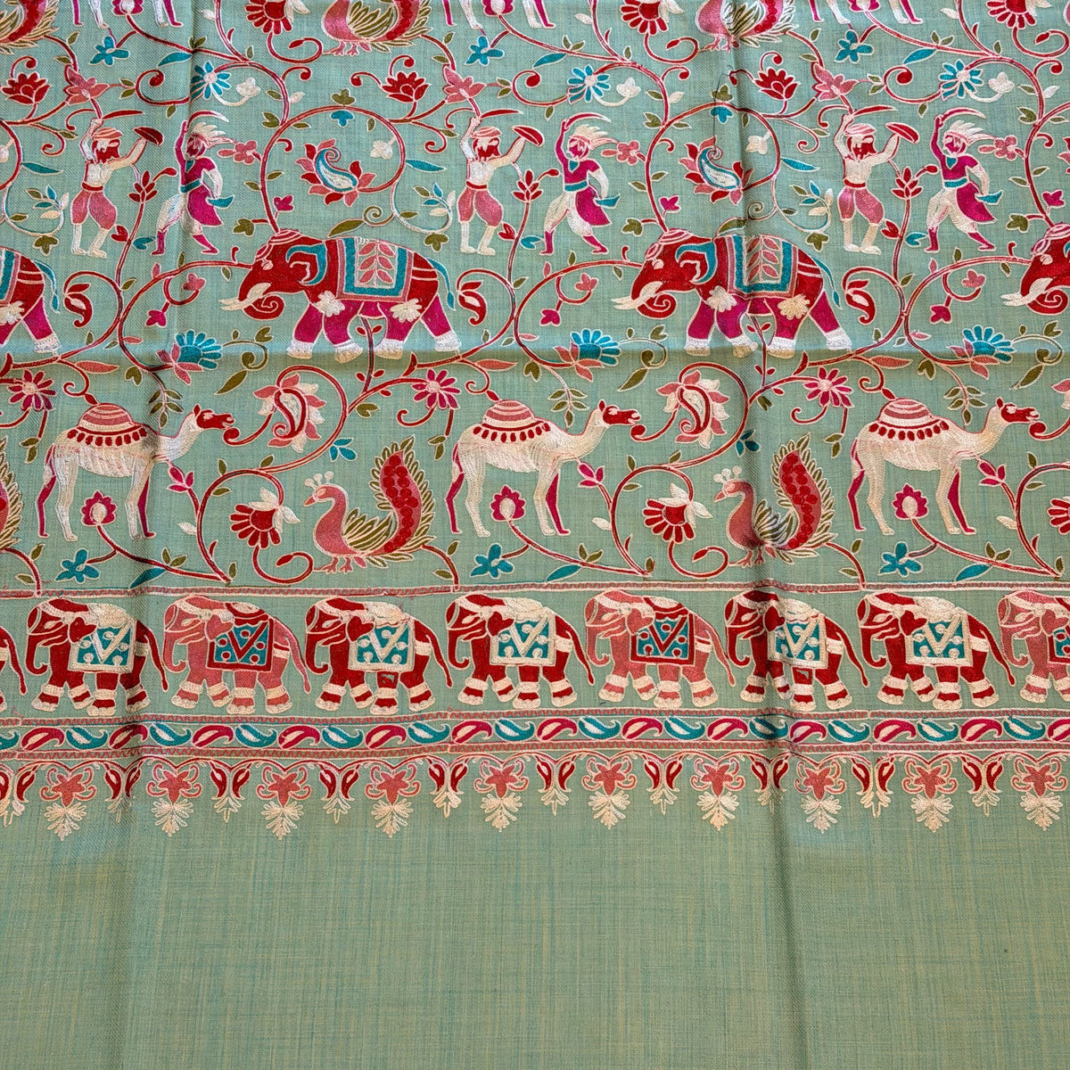 A close-up of a light sage green fabric, likely silk or satin, featuring intricate hand-woven or printed motifs in red, pink, and blue, with a decorative border on the right side.