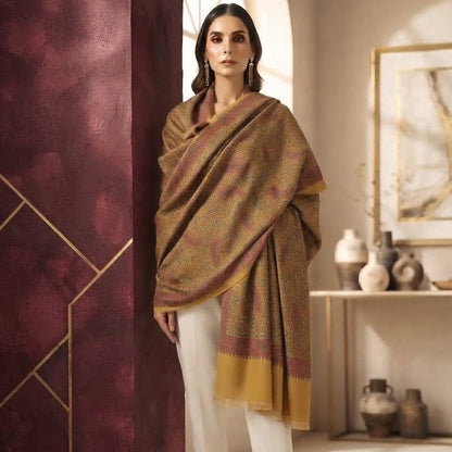 A woman models a traditional shawl in a warm brown and gold patterned fabric with a yellow border, paired with white trousers, standing in a styled interior setting.