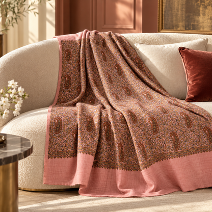 A draped throw blanket with a brown and pink paisley pattern and solid pink borders, styled on a cream-colored sofa in a warm, elegant room.