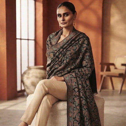 A woman seated on a cushion in a warm indoor setting, wearing a dark patterned shawl over a beige outfit with beige heels.
