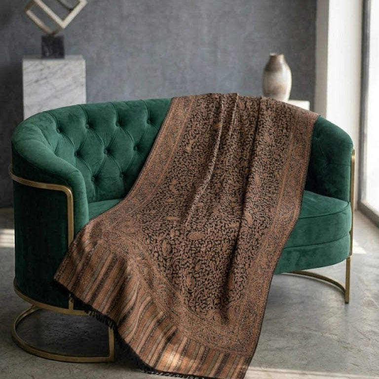 A brown patterned throw blanket with fringe and striped borders draped over a green velvet tufted armchair in a modern room setting.