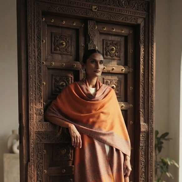A woman stands in front of an ornate carved wooden door wearing a draped orange shawl over white pants, in a softly lit indoor setting.