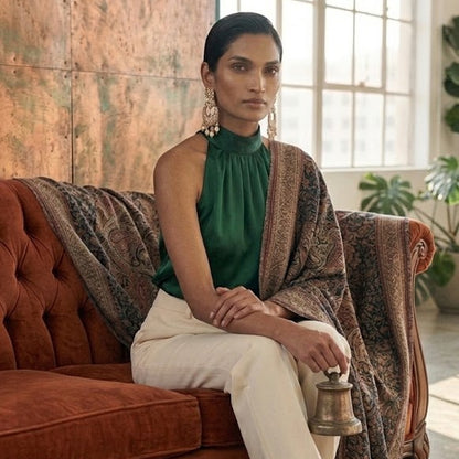 A woman sits on a rust-brown velvet sofa in a sunlit room, wearing a sleeveless dark green top, cream trousers, and a patterned shawl draped over her shoulders. She holds a small brass bell, with a large window and textured wall behind her.