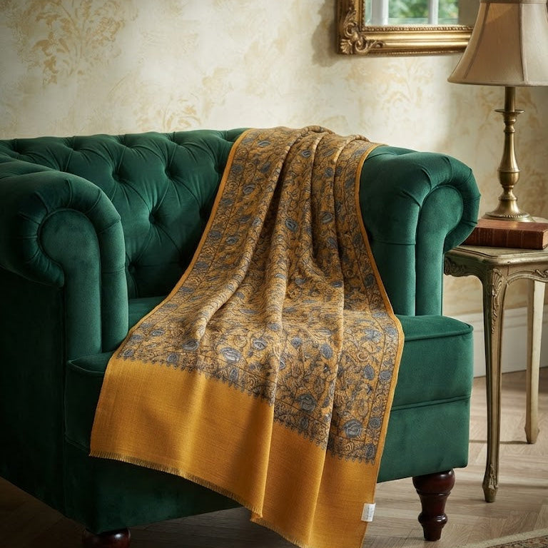 A luxurious mustard yellow and gold patterned throw blanket draped over a deep green velvet armchair in a classic, elegant room setting.