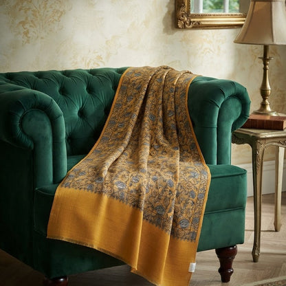 A luxurious mustard yellow and gold patterned throw blanket draped over a deep green velvet armchair in a classic, elegant room setting.