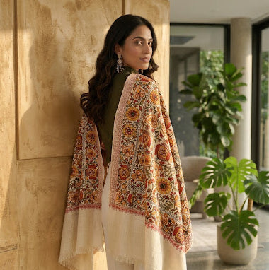 A woman wearing a cream-colored shawl with intricate orange, yellow, and red floral embroidery, draped over her shoulders, posing in a warm, sunlit indoor setting with a textured beige wall and potted plants.