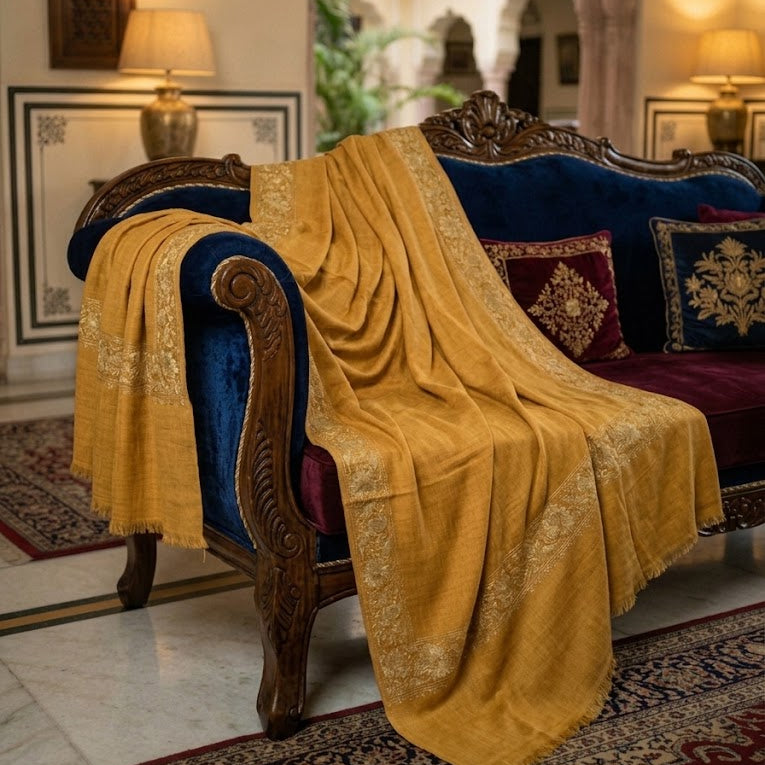 A draped mustard yellow silk throw blanket with gold embroidery is styled on an ornate dark wood and blue velvet sofa in a traditional Indian courtyard setting.