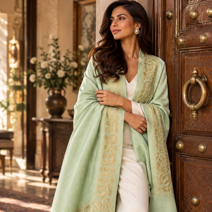 A woman stands in a doorway wearing a light sage green shawl with gold embroidered borders over a white outfit. The setting features a rustic wooden door and warm interior lighting.