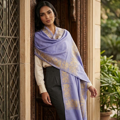 A woman wearing a lavender purple shawl with gold zari embroidery draped over her shoulders, paired with a cream long-sleeve top and dark gray trousers, standing in an ornate carved doorway.