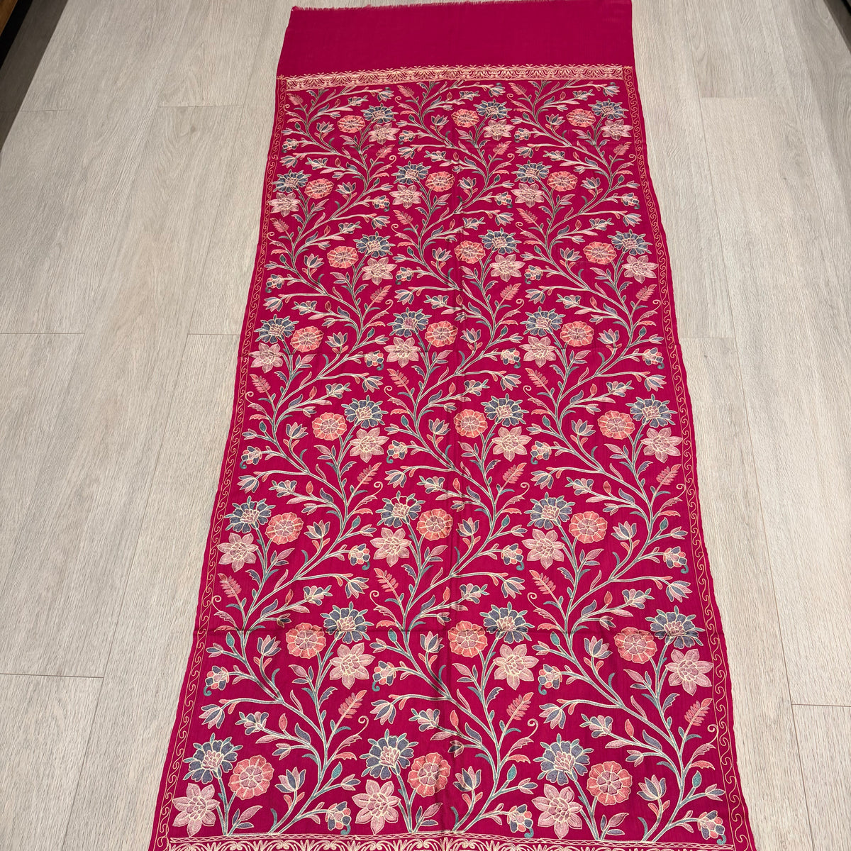 A long magenta pink embroidered table runner laid flat on a light wood floor. The runner features intricate floral and vine embroidery in pink, beige, and light blue, with solid magenta borders on both ends.