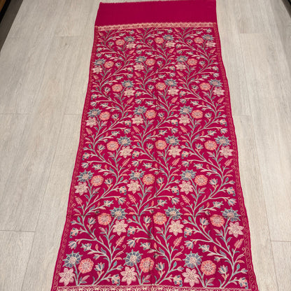 A long magenta pink embroidered table runner laid flat on a light wood floor. The runner features intricate floral and vine embroidery in pink, beige, and light blue, with solid magenta borders on both ends.