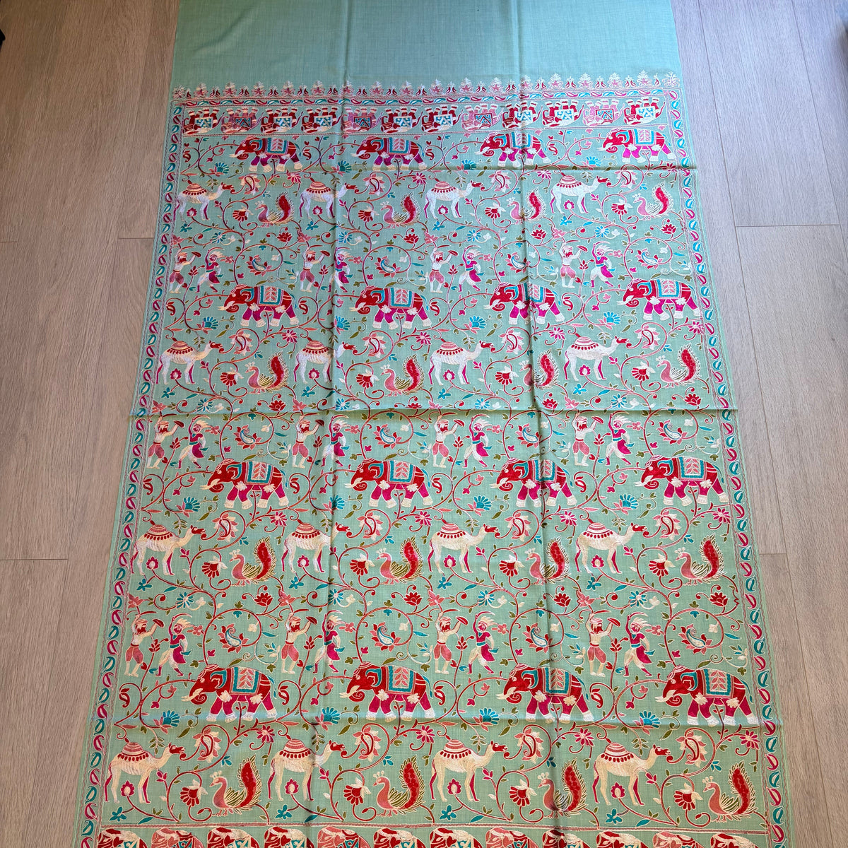 A light green Banarasi silk saree with intricate red, pink, and white embroidery depicting elephants, deer, and floral motifs, laid flat on a wooden floor.