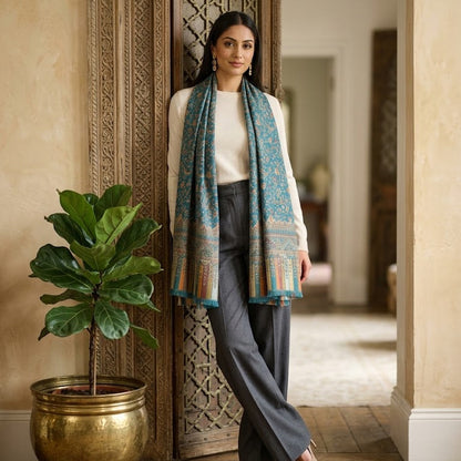 A woman stands in a doorway wearing a long patterned teal and orange scarf draped over her shoulders, a cream top, and dark gray wide-leg pants. A large green potted plant in a gold pot sits to her left in a warmly lit interior with beige walls and carved wooden door details.