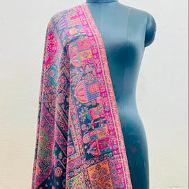 A multicolor patterned shawl with a black base and intricate pink, orange, and green traditional motifs, draped over a black mannequin torso against a plain beige wall.
