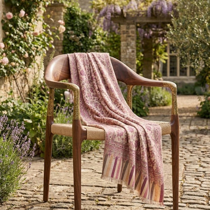 A pink patterned knit throw blanket with fringe is draped over a wooden armchair in a garden courtyard setting.