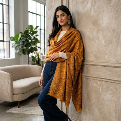 A woman wearing a mustard yellow patterned shawl draped over her shoulders, dark blue jeans, and brown ankle boots, leaning against a textured beige wall in a modern living room setting.