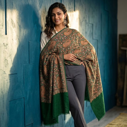 A woman models a traditional patterned shawl draped over her shoulders, paired with gray trousers and green loafers, standing against a textured blue wall.